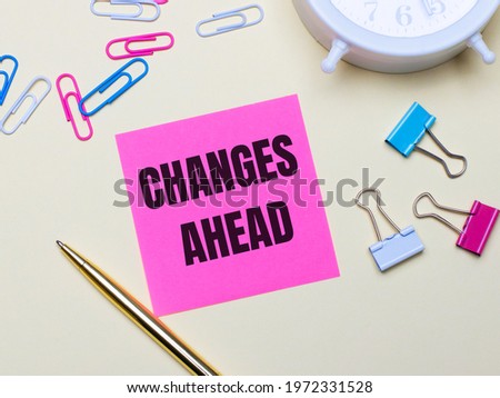 On a light background, a white alarm clock, pink, blue and white paper clips, a golden pen and a pink sticker with the text CHANGES AHEAD
