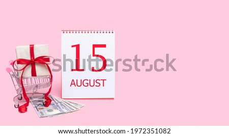 15th day of august. A gift box in a shopping trolley, dollars and a calendar with the date of 15 august on a pink background. Summer month, day of the year concept.