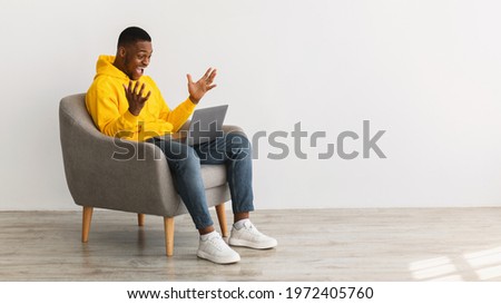 Excited African American Man With Laptop Shouting Celebrating Great Online News Sitting In Chair Over Gray Wall Indoor, Side View. Successful Freelance Career Concept. Panorama, Copy Space