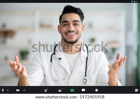 Cheerful young arab man doctor having video call with patient, giving recommendations during COVID-19 pandemic, or attending online conference while working in his cabinet in clinic or hospital