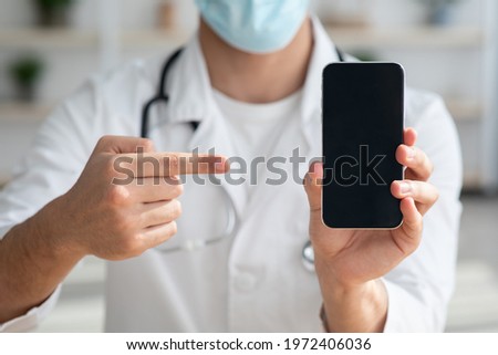 Unrecognizable doctor in face mask pointing at smartphone with empty screen, mockup, closeup. Cropped of male doc working online, recommending apps for online appointments during COVID-19 pandemic