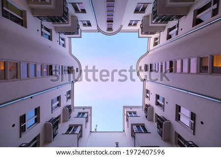 Moody symmetrical look above from backyard of an old tenement house to the roof opening with windows, walls, reflections, warm purple and blue light of the setting sun is the sky. Building well Royalty-Free Stock Photo #1972407596