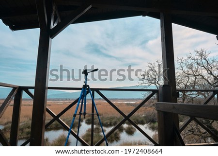 Blue color tripod for photographers standing inside bird watching tower in karacabey (longoz ormani) floodplain in bursa with mountain, dried plants and bushes, pond and lake background and blue sky