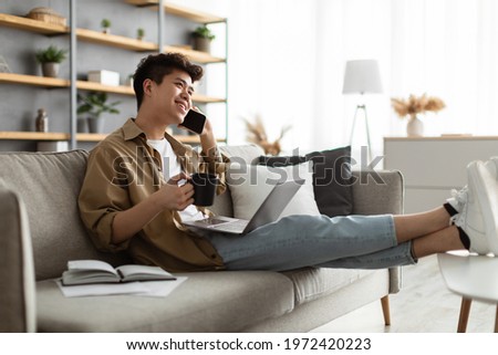 Mobile Communication. Portrait of smiling young asian guy using laptop and talking on mobile phone, holding cup and drinking coffee, working remotely sitting on the couch at home office Royalty-Free Stock Photo #1972420223