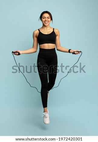 Happy fit african american lady jumping over the skipping rope, during cardio workout on blue background in studio, crop. Full length. Weight loss, active lifestyle concept Royalty-Free Stock Photo #1972420301