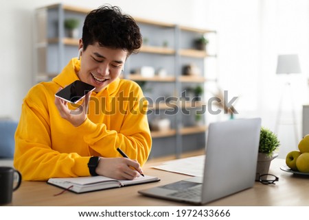 Work From Home. Young Asian Guy Talking On Mobile Phone And Taking Notes, Using Voice Assistant, Writing In Notebook. Smiling Man Sitting At Desk With Laptop At Home Office, Wearing Earbuds Royalty-Free Stock Photo #1972433666
