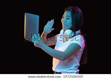 Greeting, videocall, chat. Asian young woman's portrait on dark studio background in neon light. Beautiful female model with tablet. Concept of human emotions, facial expression, youth, sales, ad.