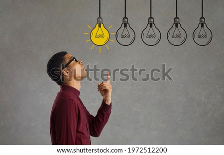 Side profile view young man making choice looking up at several electric light bulbs. Creative Afro American entrepreneur considers different effective business ideas, points finger and picks best one Royalty-Free Stock Photo #1972512200
