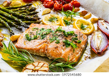 Sheet pan dinner - roasted salmon steak with asparagus, lemon ,rosemary, tomatoes, onion and garlic on cooking pan on wooden table  Royalty-Free Stock Photo #1972545680
