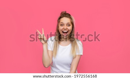Banner. A young pretty caucasian impressed excited smiling cheerful blonde woman in a white t-shirt shows a peace gesture with hand isolated on a bright color pink background.Girl shows a victory sign
