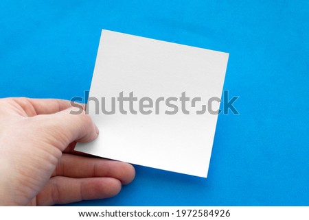 Male hand holding blank sheet of white note paper on blue background.