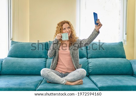 Happy redhead girl taking selfie with smartphone and drinking coffee. Young woman having fun sitting on the sofa at home. High quality photo