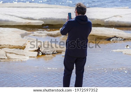 A middle-aged man in sun glasses stands on the Baltic Sea  shore on the sunny day on early April. There is ice on the water. Man is taking photo of the icy bay, using camera in his smartphone