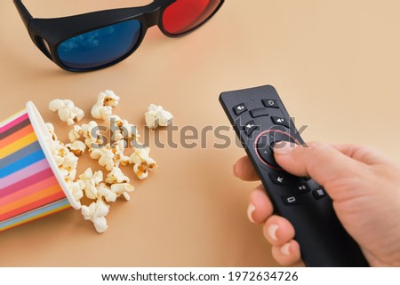 Hand holds TV remote, bucket of popcorn and 3D glasses. Flat lay, Top view. Concept pastime, entertainment and movie night watching.