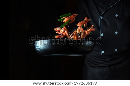 Professional chef cooks shrimp in a pan with vegetables. Cooking seafood, healthy vegetarian food and food on a dark background. Freezing in motion. Free advertising space