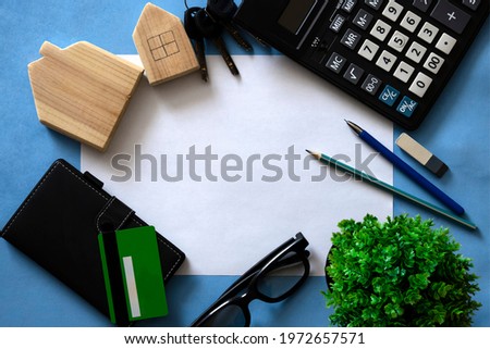 Flat lay with white sheet paper with center on blue background with wooden houses, calculator, notebook, credit card, pen, glasses and green plant. Concept construction, budgeting, property insurance.