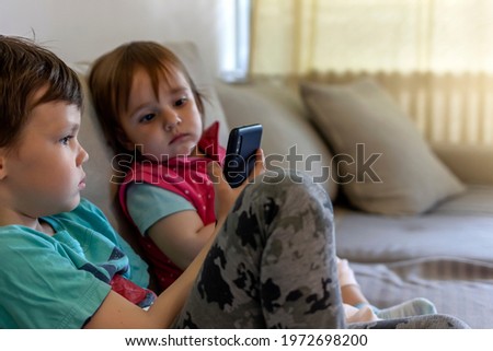 Happy little Caucasian kids sit on couch play game on smartphone together, smiling small brother and sister have fun hold using cellphone, watch funny cartoon. Children and technology concept.
