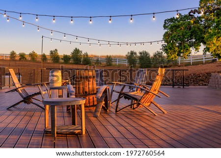 Outdoor al fresco chairs and table on a wooden deck at sunset in the spring with grape vines and hills in the background, Napa Valley, California USA Royalty-Free Stock Photo #1972760564