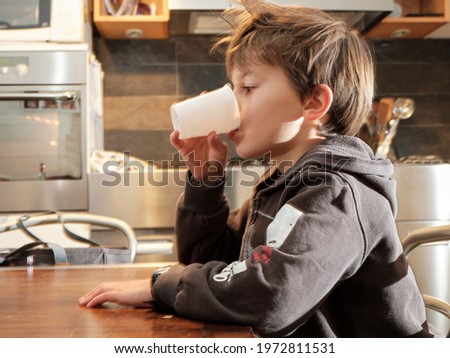 The child drinks fresh water from a white plastic cup in the home kitchen.