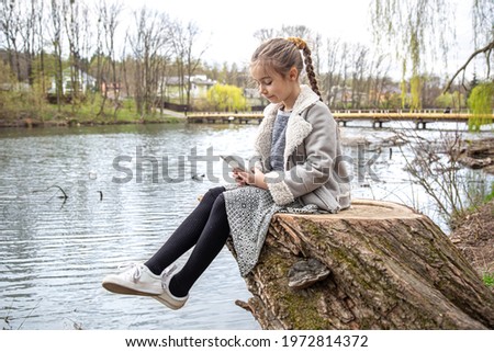 A little girl checks her phone, sitting by the river, and does not pay attention to the nature around.