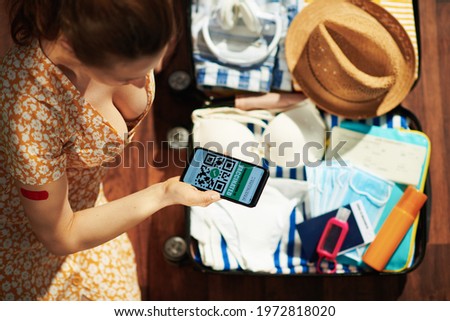 Young woman with smartphone, covid passport app, sanitizer, trolley bag, air tickets, medical mask, headphones, straw hat, spf, plaster after covid vaccine and bikini in the modern living room.