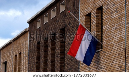 Dutch national flag hanging half mast on the exterior facade of a modern building in commemoration of those fallen in war times with blue sky and clouds in the background