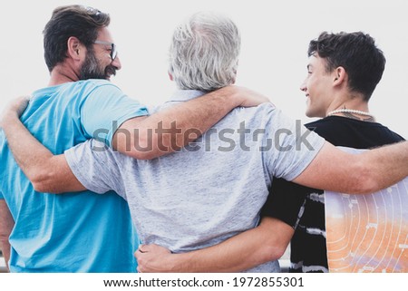 Multigenerational family group standing outdoors by the sea, hugging each other and smiling. Grandfather, son and teenage grandson. Royalty-Free Stock Photo #1972855301