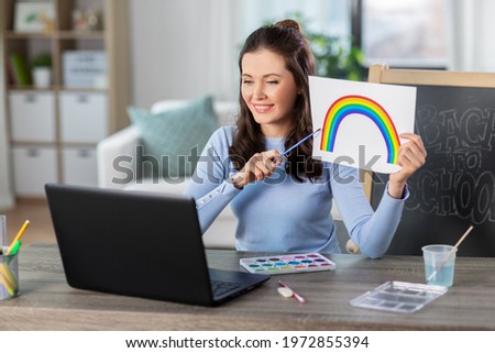 distant education, school and people concept - happy smiling female teacher with laptop computer and picture of rainbow and colors having online art class at home