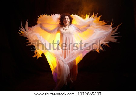 Brunette girl in an elegant dress and with white angel wings on a black background. Model, actress or dancer posing in the studio