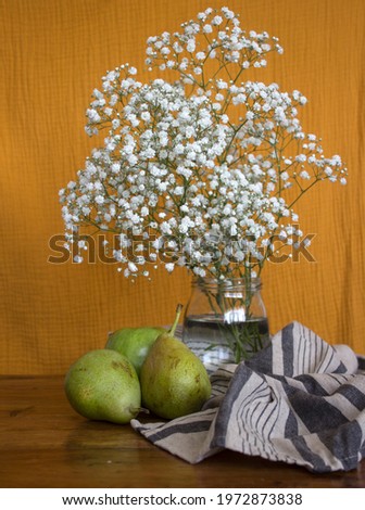 Two green pears, apple and white flowers in glass vase on wooden table. Still life with fruits and flowers. Yellow textured background. 