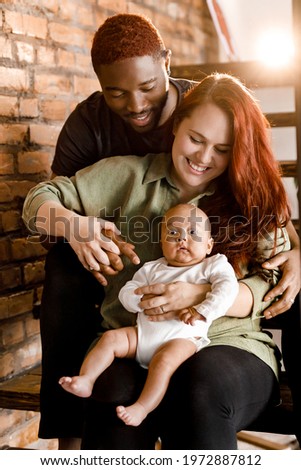 Portrait of mixed race smiling family sit on the stairs. Beautiful mother and smiling father holding baby boy. Joyful parents looking gently at their son spend weekends at home, childcare concept