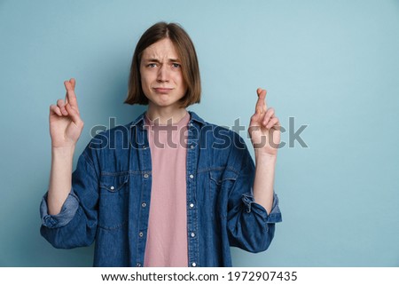 Young man frowning while holding fingers crossed for good luck isolated over blue background