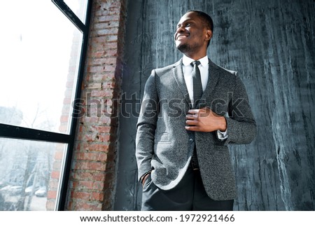 Handsome smiling african american manager in a suit buttons his jacket and looks to the side while standing against the background of the window Royalty-Free Stock Photo #1972921466