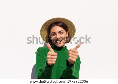 Stylish woman in green casual sweater and hat outdoor on white wall background cheerful, joyful and excited enjoys hot summer day, positive vibes only, show thumbs up Royalty-Free Stock Photo #1972922306
