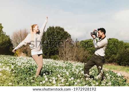 Young man taking a pictures of his girlfriend standing outdoors with arms raised. Couple on camp taking photographs with digital camera.