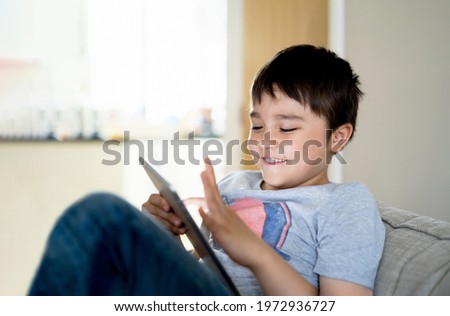 Authentic Kid sitting on sofa watching cartoons or playing games on tablet,Child boy using digital pad learning lesson online on internet,Home schooling,Distance learning online education concept