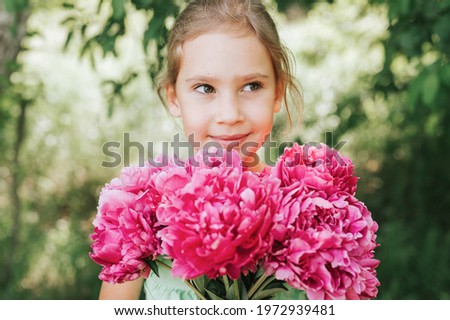 portrait of a happy cute little caucasian seven year old kid girl, holds in hands a bouquet of pink peony flowers in full bloom on the green background of nature