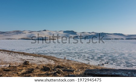 Snow patterns and several minibuses are visible on the surface of the frozen lake. Snow-capped mountain range against the blue sky. In the foreground there are picturesque stones and dry grass. Baikal
