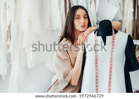 Female fashion designer posing in her creative workshop. Young woman business enterpreneur looking to the camera with mannequin during preparing garment for a new collection in her creative office. Royalty-Free Stock Photo #1972946729