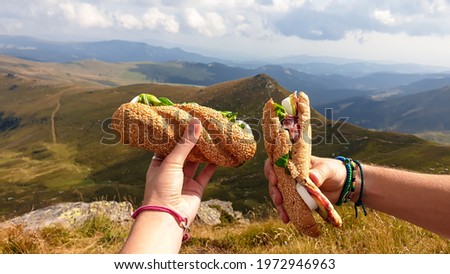 Lunch break in Austrian Alps. Two hands holding sandwiches with the view on high Alps. There are endless mountain chains in the back. Eating in the fresh air. A bit of overcast Royalty-Free Stock Photo #1972946963