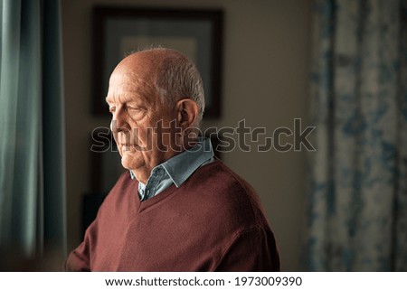 Lonely old man in contemplation near window looking away and thinking. Thoughtful retired man at home looking outside window and contemplating. Pensive senior with sorrow and distressed expression. Royalty-Free Stock Photo #1973009390