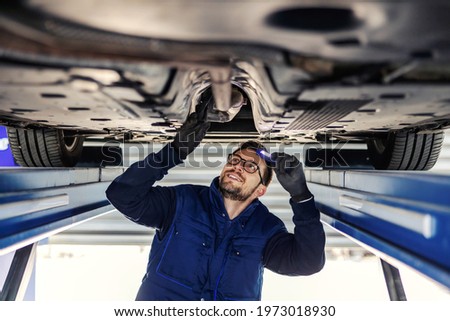 Technical inspection of the car. Car service in the workshop. A man in a blue uniform stands under a car in the garage and checks the car’s axles. It illuminates the chassis with a flashlight Royalty-Free Stock Photo #1973018930