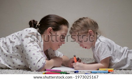 Happy family playing together at home on floor. Child and Mom, nanny, teaches girl to draw. A mother helps her daughter learn to draw on paper, coloring with multi-colored pencils and felt-tip pens.