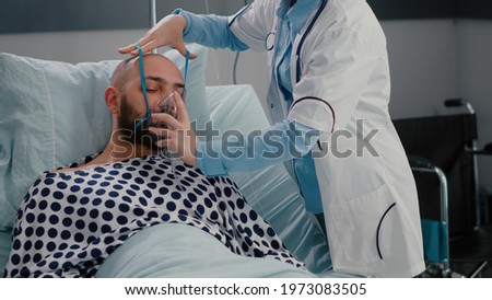 Patient sitting in bed while woman doctor putting oxygen mask monitoring respiratory illness. Physician medic writing sickness treatment working in hospital ward during recovery appointment