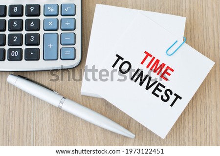 TIME TO INVEST is written on a white note card on a wooden background near the calculator and pen. Investment concept