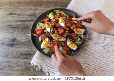 Nicoise salad, beans, canned tuna, tomatoes, eggs, lettuce on a dark plate on a wooden table. Female hands wrestle with a fork and knife over the salad. Horizontal top view.