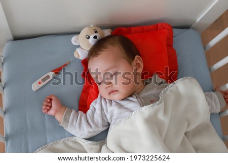 Baby boy sick sleep in cot next to digital thermometer. Mixed race Asian-German infant sickness, fever lying on bed. Royalty-Free Stock Photo #1973225624