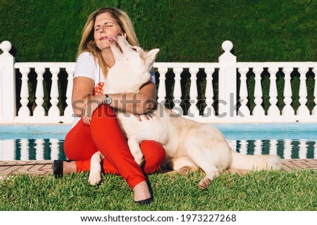 Mature blonde woman seated on the grass with siberian husky dog licking face near to the swimming pool