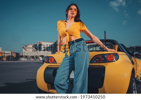 Female in blue jeans, orange top and sunglasses is posing near yellow car roadster with paper cup of tea on trunk at parking lot. Copy space Royalty-Free Stock Photo #1973410250