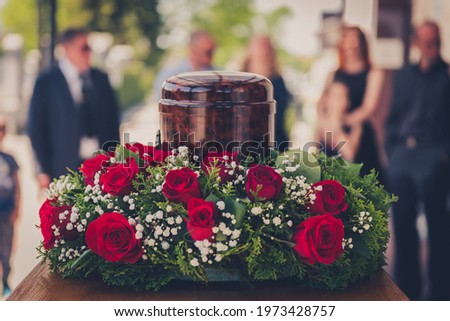 Funerary urn with ashes of dead and flowers at funeral. Burial urn decorated with flowers and people mourning in background at memorial service, sad and grieving last farewell to deceased person. Royalty-Free Stock Photo #1973428757